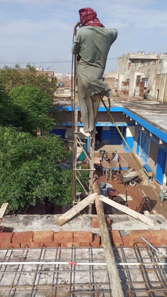 Picture of a construction worker balancing on the roof of the new building. Picture shows the other part of the school building and the school yard as well as surrounding buildings.